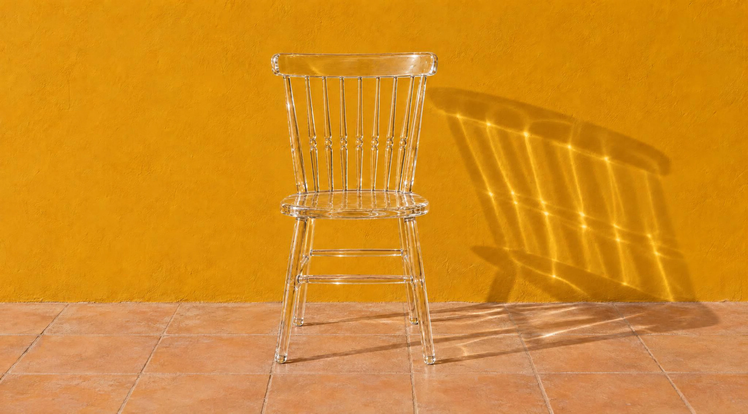 Clear glass spindle-back chair in front of an ochre wall, its shadow filled with bright lines of refracted sunlight. Made with GPT Image 2.5 Sunburst.