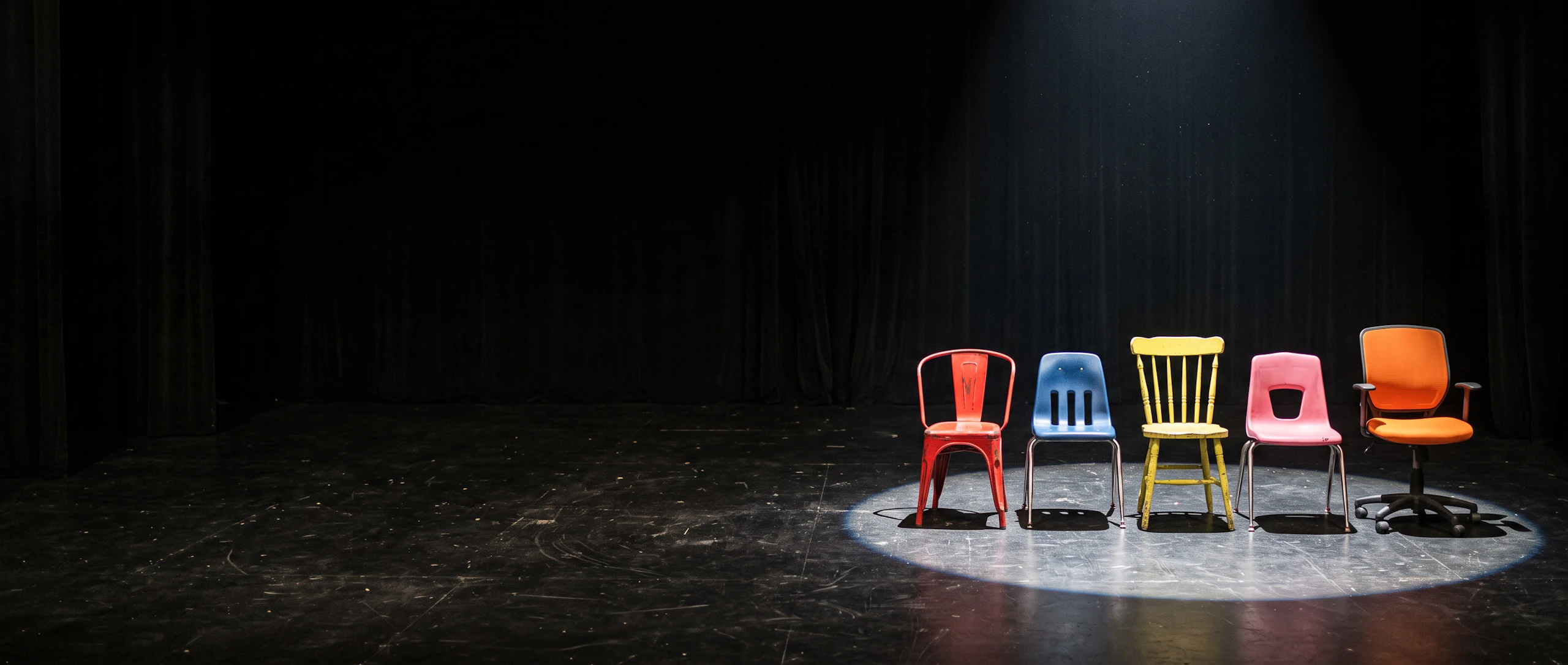 Five mismatched chairs in red, blue, yellow, pink and orange wait in one spotlight on a dark stage. Made with Nano Banana 2.1.