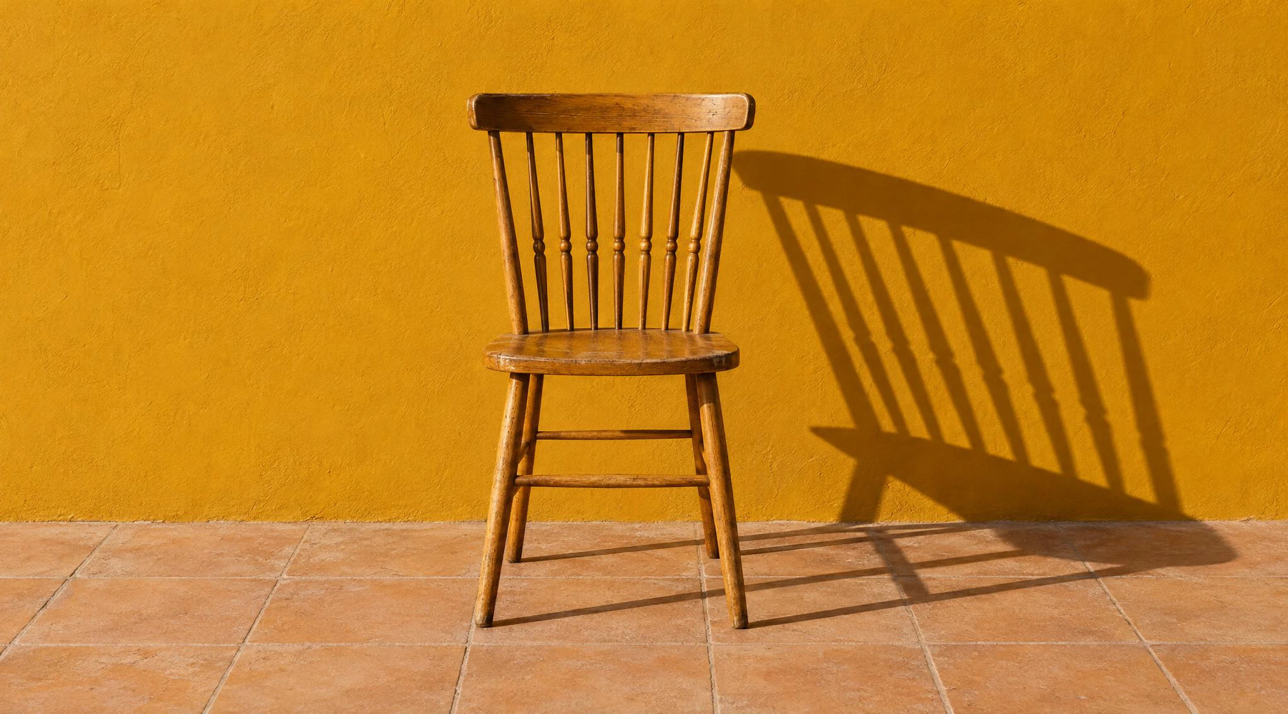 Wooden spindle-back chair in front of an ochre wall, casting a long hard shadow in low sun. Made with GPT Image 2.5 Flare.