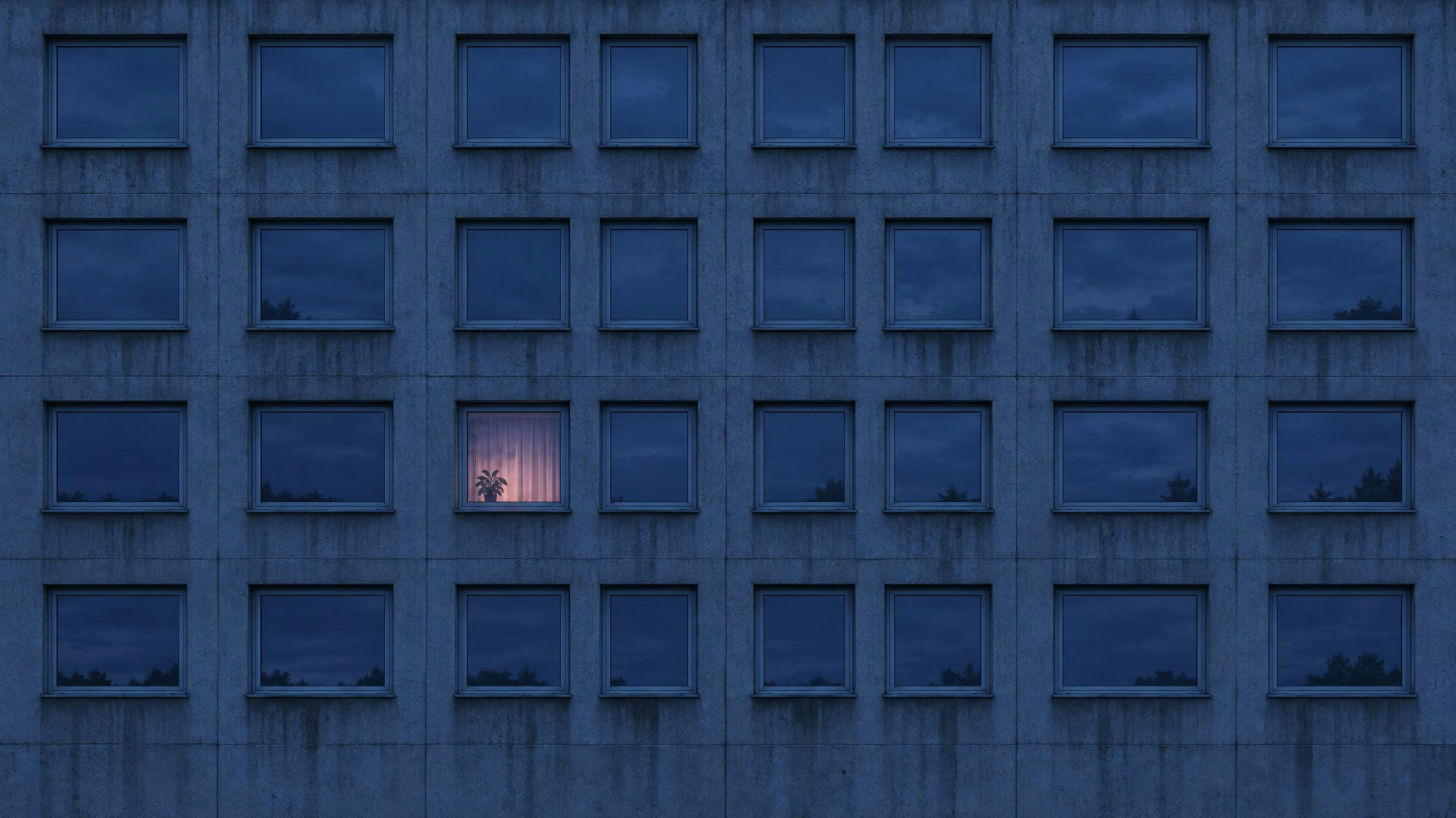 A concrete tower block at dusk, every window dark except one glowing pink behind a curtain, with a plant on the sill. Made with GPT Image 2.5 Flare.