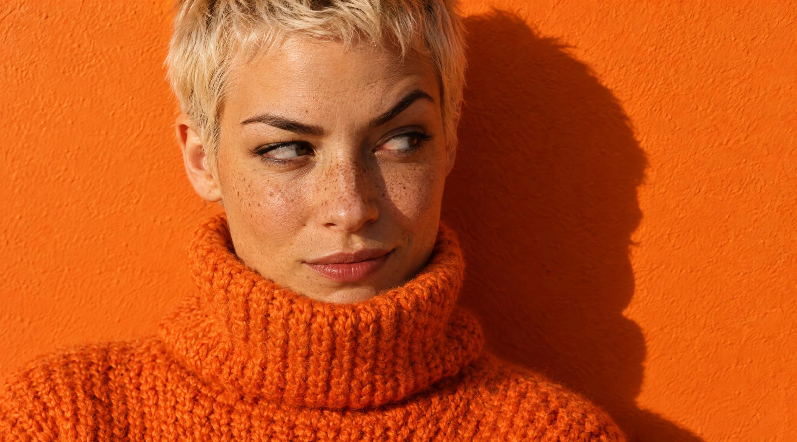 Photo of a freckled woman with a bleached-blonde crop in a tangerine roll-neck, glancing sideways against a tangerine wall in hard sunlight. Made with GPT Image 2.5 Flare.